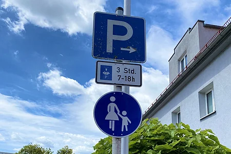 Verkehrsschilder an Laterne: Parken rechts 3 Stunden von 7 bis 18 Uhr, darunter Fußgängerzone mit Frau und Kind.