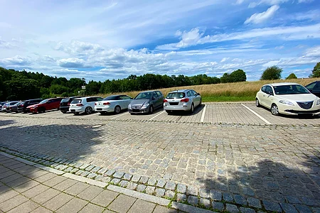 Parkplatz mit mehreren geparkten Autos, im Hintergrund Wiese und Bäume unter blauem Himmel mit Wolken