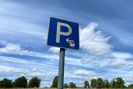 Blaues Parkplatzschild mit weißem P und Pfeil nach links vor Wiese und bewölktem Himmel