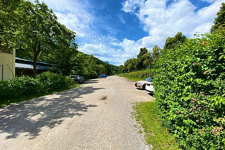 Breiter Kiesweg mit parkenden Autos rechts und Bäumen links unter blauem Himmel mit Wolken.