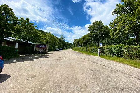 Breite Schotterstraße mit parkenden Autos, Bäumen und blauem Himmel mit Wolken an einem sonnigen Tag.
