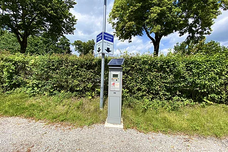 Parkscheinautomat mit Solarpanel vor Hecke und Bäumen an einem Kiesweg unter bewölktem Himmel.