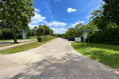 Parkplatz mit Schotterboden, Bäumen und blauem Himmel, zwei Parkschilder links und rechts sichtbar