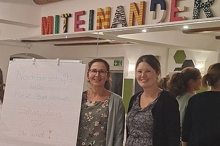 Zwei Frauen stehen neben einem Flipchart mit dem Text „Nachbarschaftshilfe Markt Bad Abbach Schön, dass SIE da sind!“ vor einem Spiegel mit bunten Buchstaben „MITEINANDER“.