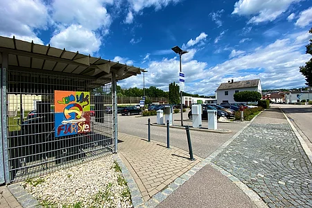 Parkplatz mit Schrankenanlage und buntem Schild an einem Zaun, im Hintergrund Häuser und bewölkter Himmel.