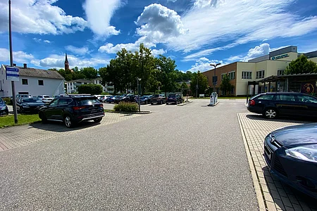 Parkplatz mit mehreren geparkten Autos, Bäumen und Gebäuden unter blauem Himmel mit Wolken