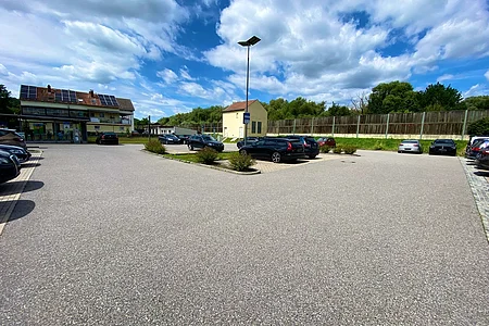 Parkplatz mit mehreren geparkten Autos, einem Haus mit Solarpaneelen und bewölktem Himmel im Hintergrund