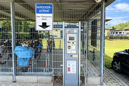 Parkscheinautomat vor Fahrradunterstand mit blauem Mülleimer und Autos im Hintergrund