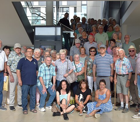 Große Gruppe von älteren und mittleren Erwachsenen steht und sitzt in einem modernen Innenraum vor einer Treppe.