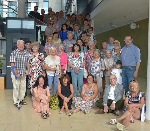 Gruppenfoto von etwa 40 Personen unterschiedlichen Alters in einem modernen Innenraum vor Treppe und Glaswand.