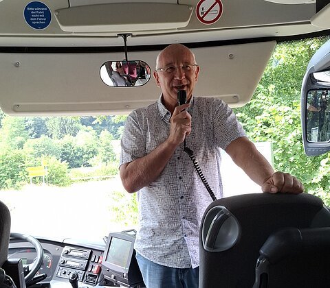 Mann spricht mit Mikrofon im Bus, im Hintergrund grüne Bäume und Verkehrsschilder sichtbar.