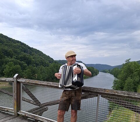 Mann mit Strohhut spielt Akkordeon auf einer Holzbrücke über einem Fluss, umgeben von grünen Hügeln.