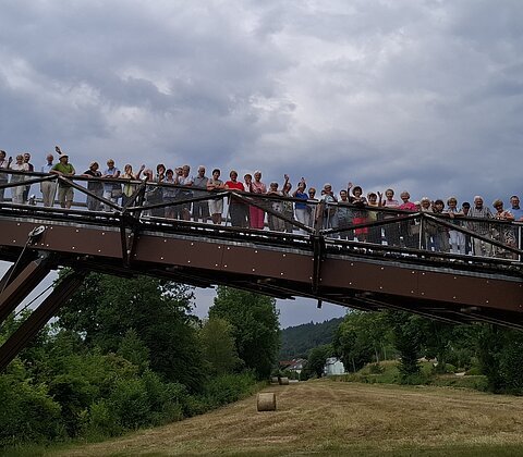 Gruppe von Menschen steht winkend auf einer Brücke über eine Wiese mit Heuballen unter bewölktem Himmel.