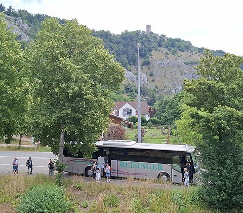 Reisebus mit der Aufschrift „Reisinger“ hält an einer Straße, umgeben von Bäumen und Bergen im Hintergrund.