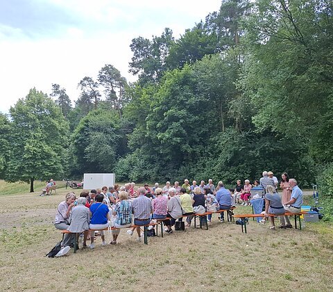 Gruppe von Menschen sitzt auf Bänken auf einer Wiese vor Wald an einem bewölkten Tag.