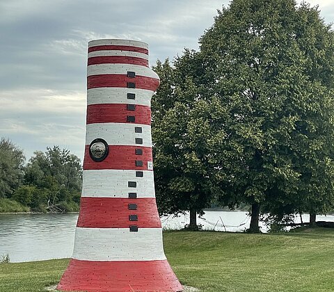 Rot-weiß gestreifter Leuchtturm aus Stein auf einer Wiese neben einem Fluss und Bäumen im Hintergrund.