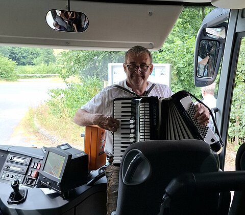 Mann spielt Akkordeon im vorderen Bereich eines Busses, Blick auf Armaturenbrett und Außenspiegel.