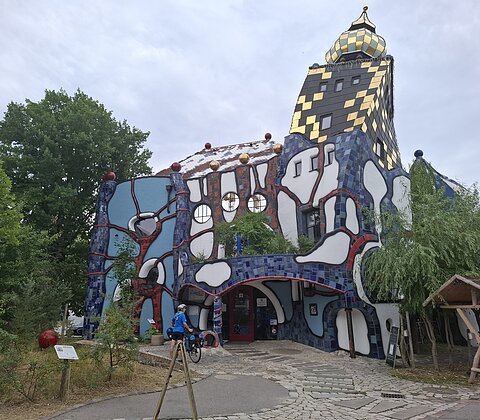 Außenansicht eines farbenfrohen, unregelmäßig geformten Gebäudes mit gold-schachbrettartigem Turm und Bäumen im Vordergrund.