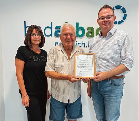 Drei Personen stehen vor einer Wand mit Schriftzug, zwei halten eine gerahmte Urkunde.