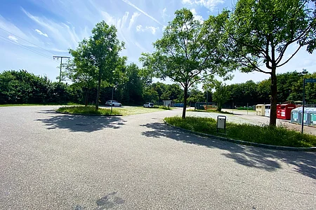 Parkplatz mit Bäumen, zwei geparkten Autos und Containern unter blauem Himmel mit Wolken