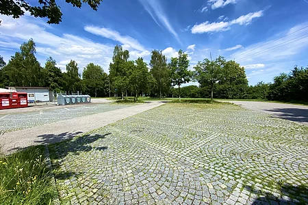 Parkanlage mit gepflastertem Platz, Bäumen, Containerstation und blauem Himmel mit Wolken.