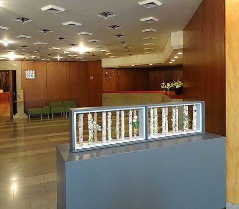 Innenraum mit Holzvertäfelung, Sitzbank, Deckenleuchten und einer Dekoration aus Birkenstämmen mit Blumen in einem Rahmen auf einem grauen Podest