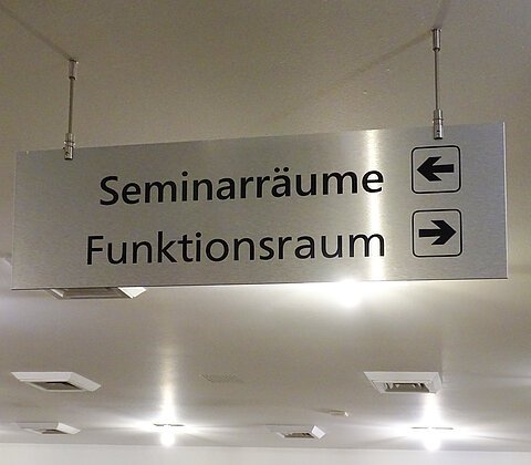 Hinweisschild an der Decke mit den Worten „Seminarräume“ und Pfeil nach links sowie „Funktionsraum“ und Pfeil nach rechts