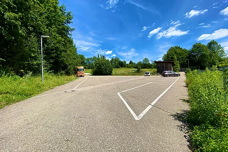 Großer Parkplatz mit zwei Autos, einem LKW, umgeben von Bäumen und blauem Himmel mit Wolken.