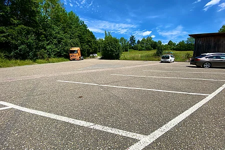Großer Parkplatz mit wenigen Fahrzeugen, umgeben von Bäumen und Wiesen unter blauem Himmel.
