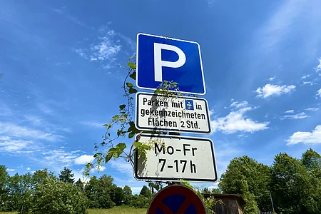 Blauer Parkplatzschild mit Zusatzschild für Parkdauer von 2 Stunden Mo-Fr 7-17 Uhr, umrankt von grünen Pflanzen.