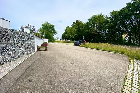 Leere asphaltierte Straße mit Steinmauer links, parkendem Auto rechts und Bäumen im Hintergrund.