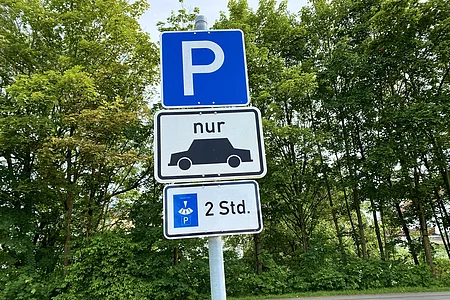 Blauer Parkplatzschild mit Zusatzschild für Pkw und Parkscheibe, Parkzeit auf 2 Stunden begrenzt, Bäume im Hintergrund.
