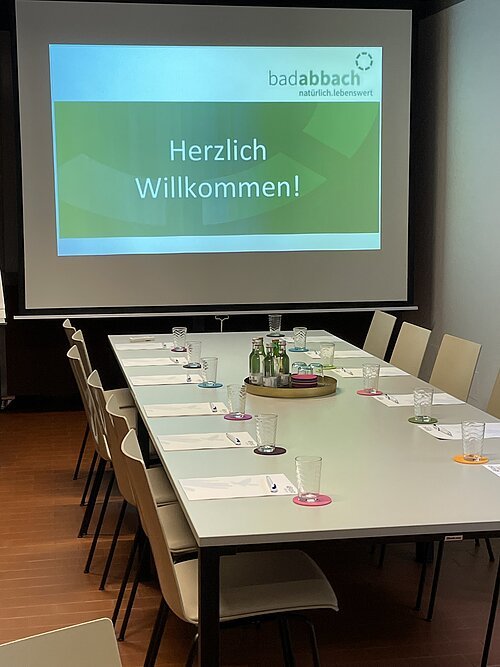 Konferenzraum mit langem Tisch, Stühlen, Gläsern, Flaschen und Projektionsbild mit Text „Herzlich Willkommen!“