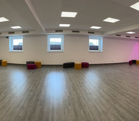 Großer leerer Raum mit Holzfußboden, weißen Wänden, Spiegelwand und bunten Sitzhockern, Deckenleuchten und lila Wandbeleuchtung.