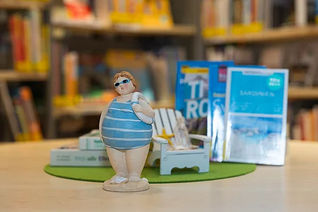 Figur einer Person mit Badeanzug und Schwimmreifen vor Büchern und Stuhl auf grünem Untergrund auf Tisch