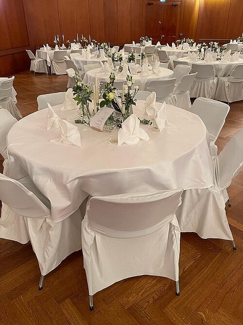 Rund gedeckter Tisch mit weißen Hussen, Servietten und Blumenarrangements in einem Saal mit Holzwänden.