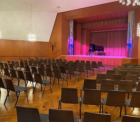 Leerer Konzertsaal mit Reihen von Stühlen und einem Flügel auf der Bühne mit roter Beleuchtung.