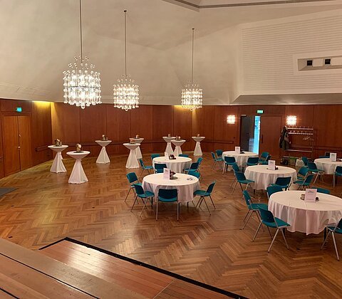 Großer Veranstaltungsraum mit runden Tischen und blauen Stühlen, Holzparkettboden und Kronleuchtern an der Decke.