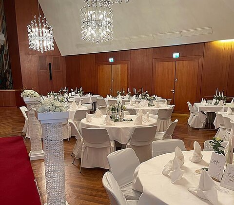 Festlich gedeckte runde Tische mit weißen Hussen und Servietten in einem Saal mit Holzverkleidung und Kronleuchtern.