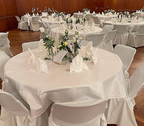 Rund gedeckter Tisch mit weißen Hussen, Servietten und Blumenarrangements in einem Saal mit Holzwänden.