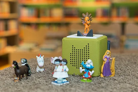 Grüner Tonie-Box Lautsprecher mit verschiedenen kleinen Spielfiguren auf Teppich vor unscharfem Bücherregal.