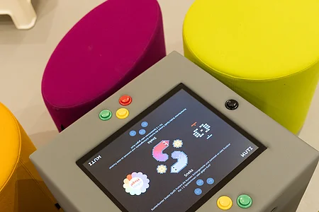 Grauer Spieltisch mit Bildschirm und bunten Knöpfen, umgeben von runden Sitzhockern in Gelb und Magenta.