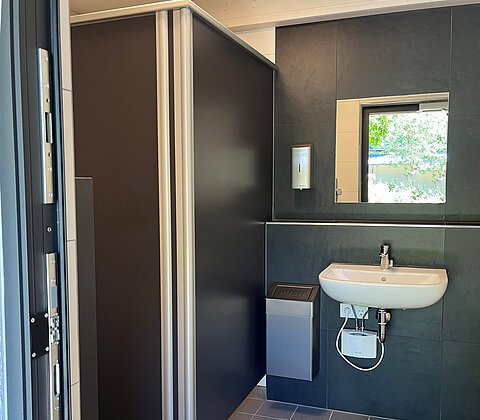 Barrierefreies WC mit grauen Trennwänden, Waschbecken, Spiegel, Seifenspender und Mülleimer.