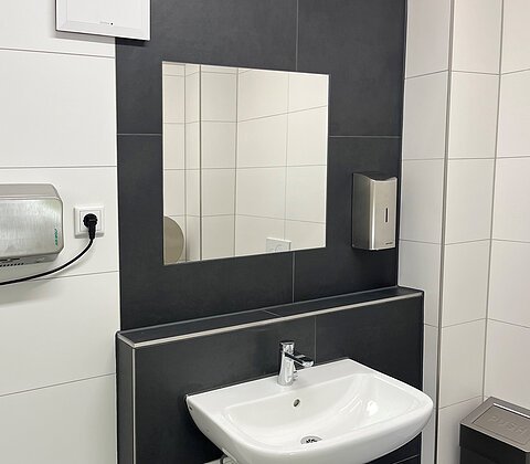 Waschbecken mit Spiegel, Seifenspender und Handtrockner in einem modernen, schwarz-weiß gefliesten Badezimmer.