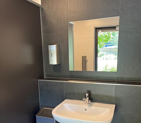 Waschbecken mit Spiegel, Seifenspender und Mülleimer in einem modernen, dunklen Badezimmer mit Fensterblick.