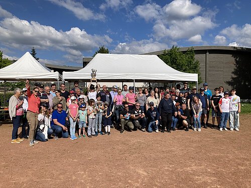 Große Gruppe von Menschen unterschiedlichen Alters vor zwei weißen Zelten auf Sportplatz bei sonnigem Wetter, eine Person hält Pokal hoch.