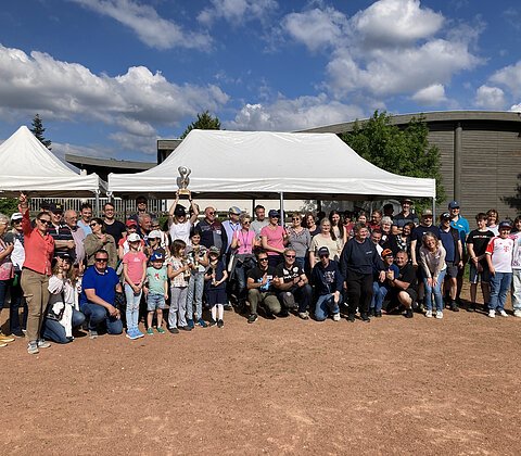 Große Gruppe von Menschen unterschiedlichen Alters vor zwei weißen Zelten auf Sportplatz bei sonnigem Wetter, eine Person hält Pokal hoch.