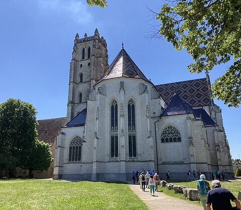 Gotische Kirche mit Turm, Menschen auf Weg davor, grüner Rasen und Bäume bei blauem Himmel