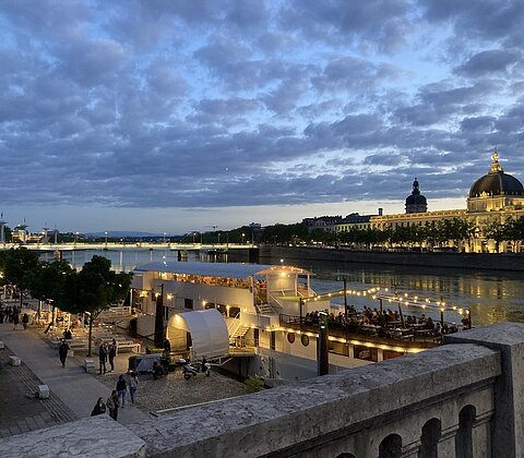 Abendliche Flussuferpromenade mit beleuchtetem Partyschiff und historischer Kuppelgebäude im Hintergrund.
