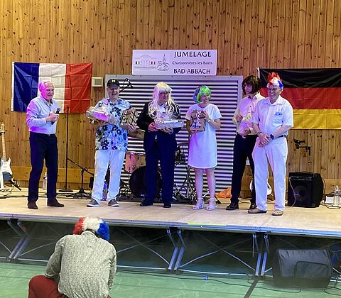 Sechs Personen stehen auf einer Bühne vor französischer und deutscher Flagge, einige tragen bunte Perücken, eine Person fotografiert.